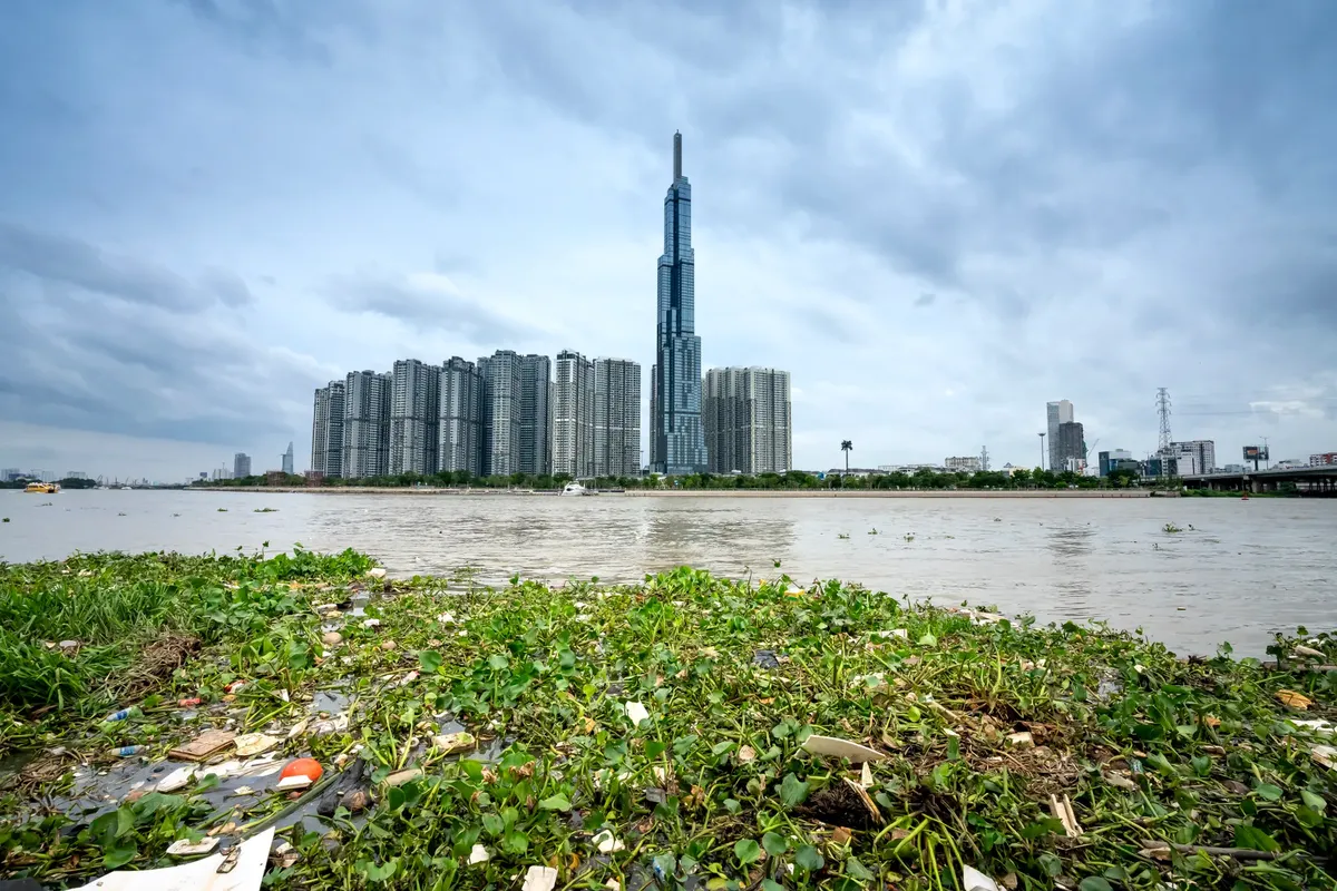 Grassy coast covered in garbage near water and of Ho Chi Minh City in Vietnam with skyscrapers near Landmark 81 building near trees in summer day under cloudy s