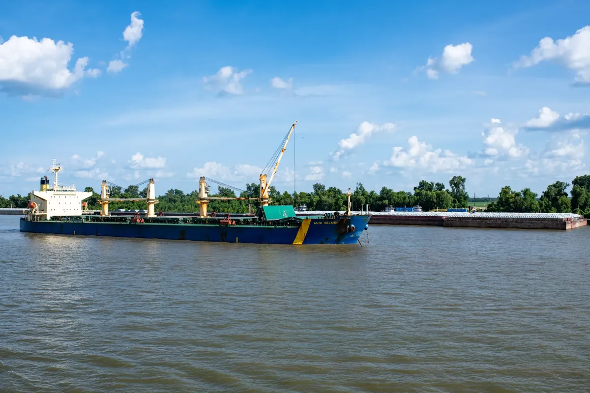 A cargo ship navigates the Mekong River surrounded by a lush green forest under a clear blue sky.