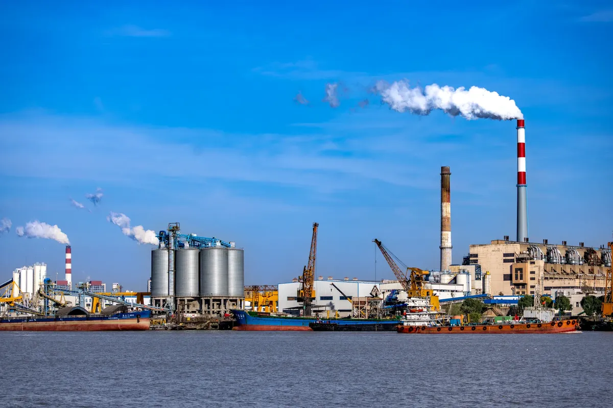 An industrial port scene with smokestacks, silos, and cranes by the water, showcasing urban industry.