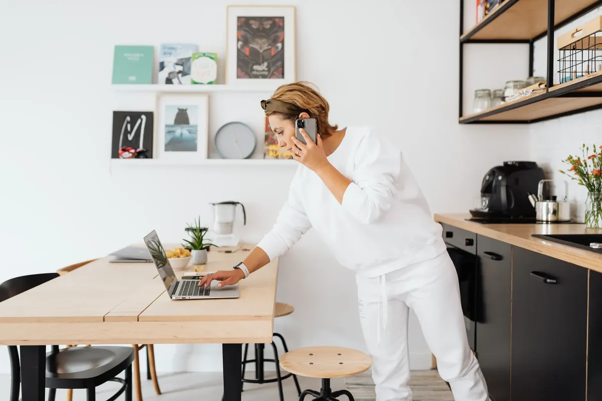 Woman using laptop and phone in stylish kitchen, blending work and home life.