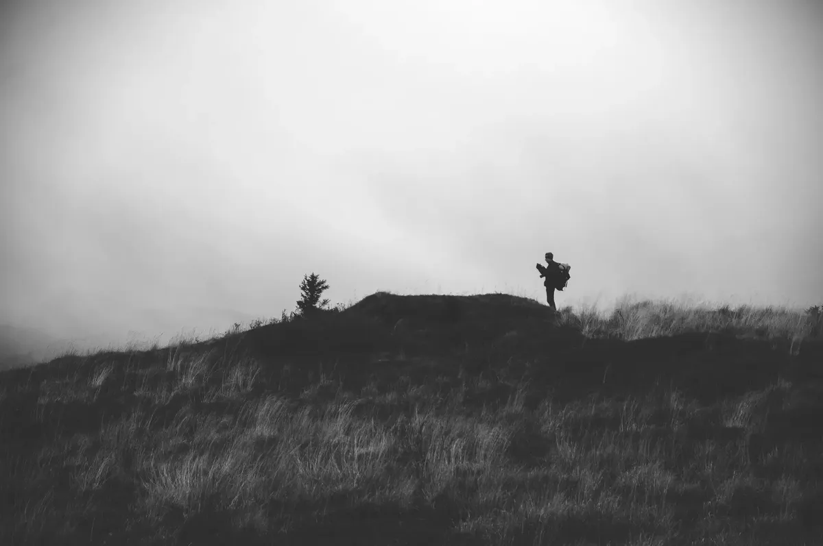 Black and white side view of distant unrecognizable person standing on grassy hill against foggy sky during hiking in nature
