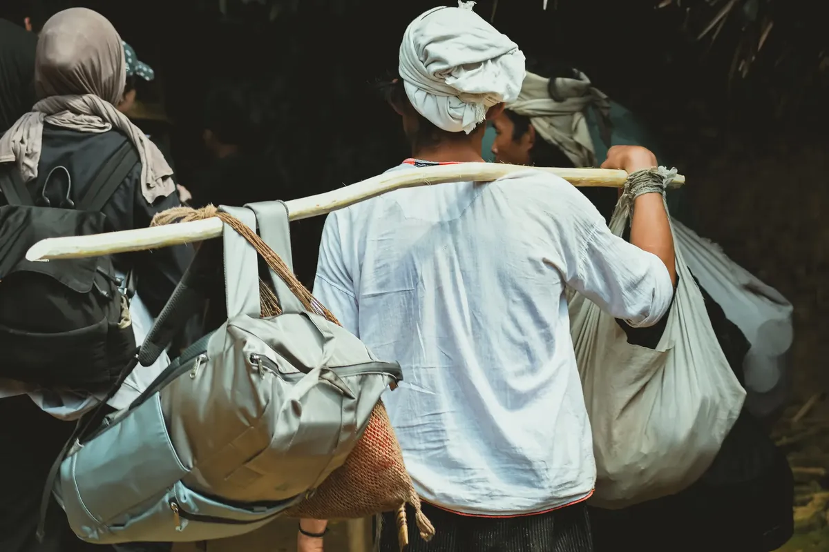 A traveler carrying multiple bags using a wooden pole, going through a rural setting.
